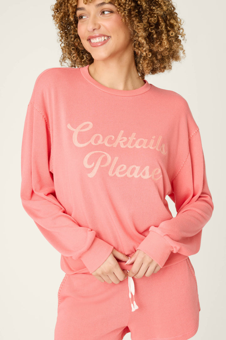 A person with curly hair wears the Paloma Mama Cocktails Please Graphic Sweatshirt by P.J. Salvage—a cozy pink French terry top with light script—while smiling and looking to the side, paired with matching shorts.