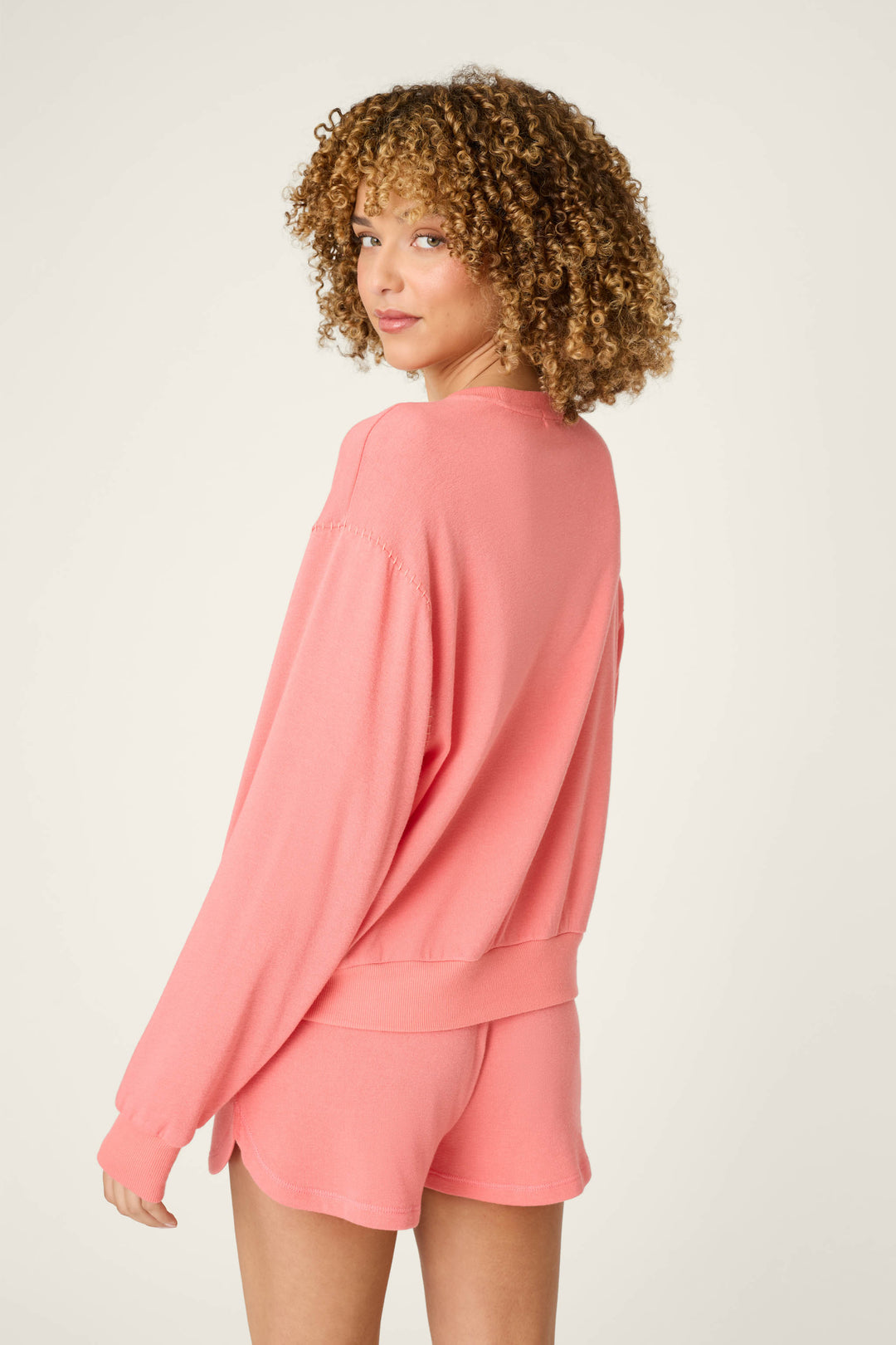 A person with curly hair wears the P.J. Salvage Paloma Mama Cocktails Please Graphic Sweatshirt, styled with matching pink shorts, standing against a light background and looking over their shoulder at the camera.