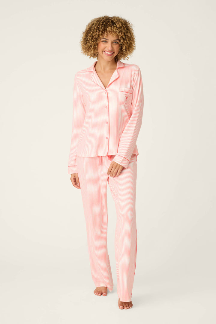 A person stands barefoot against a plain background, smiling in the Paloma Mama Sweet Stripes Long 3-Piece PJ Set by P.J. Salvage—a grapefruit pink striped sleepwear set with long sleeves, a collar, front buttons, and long pants.