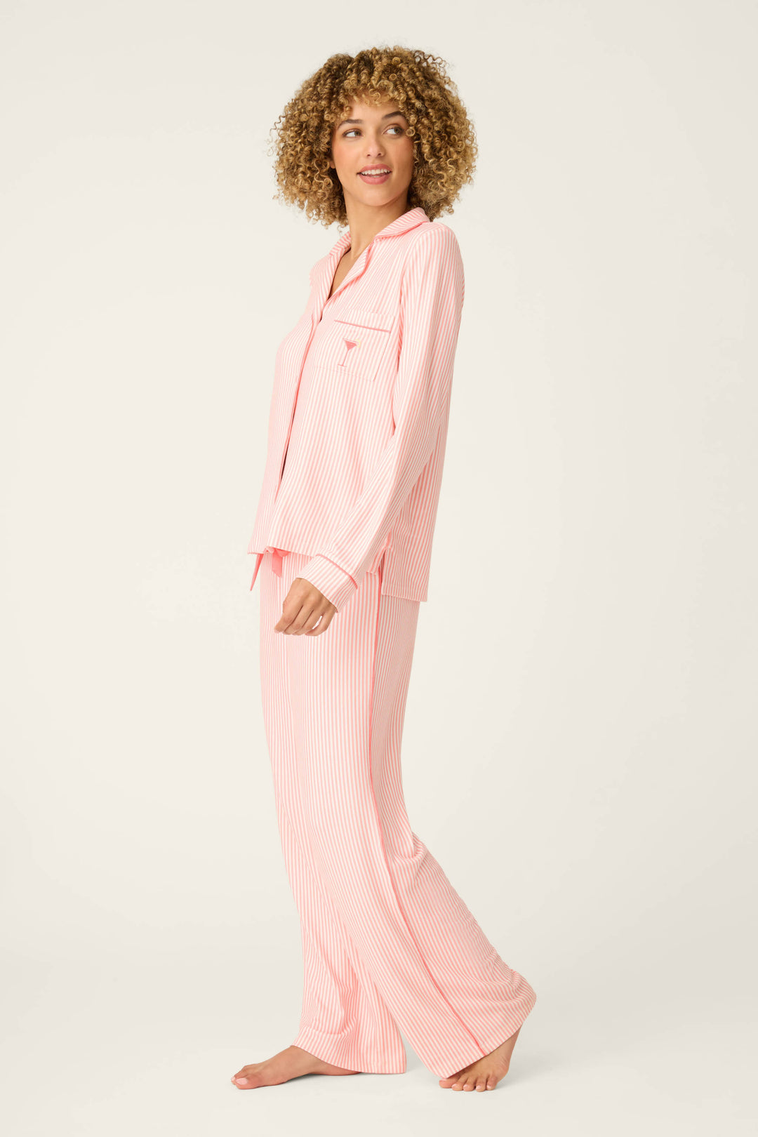 A person with curly hair stands barefoot against a plain light background, wearing the Paloma Mama Sweet Stripes Long 3-Piece PJ Set by P.J. Salvage, featuring grapefruit pink stripes and long sleeves with full-length pants.