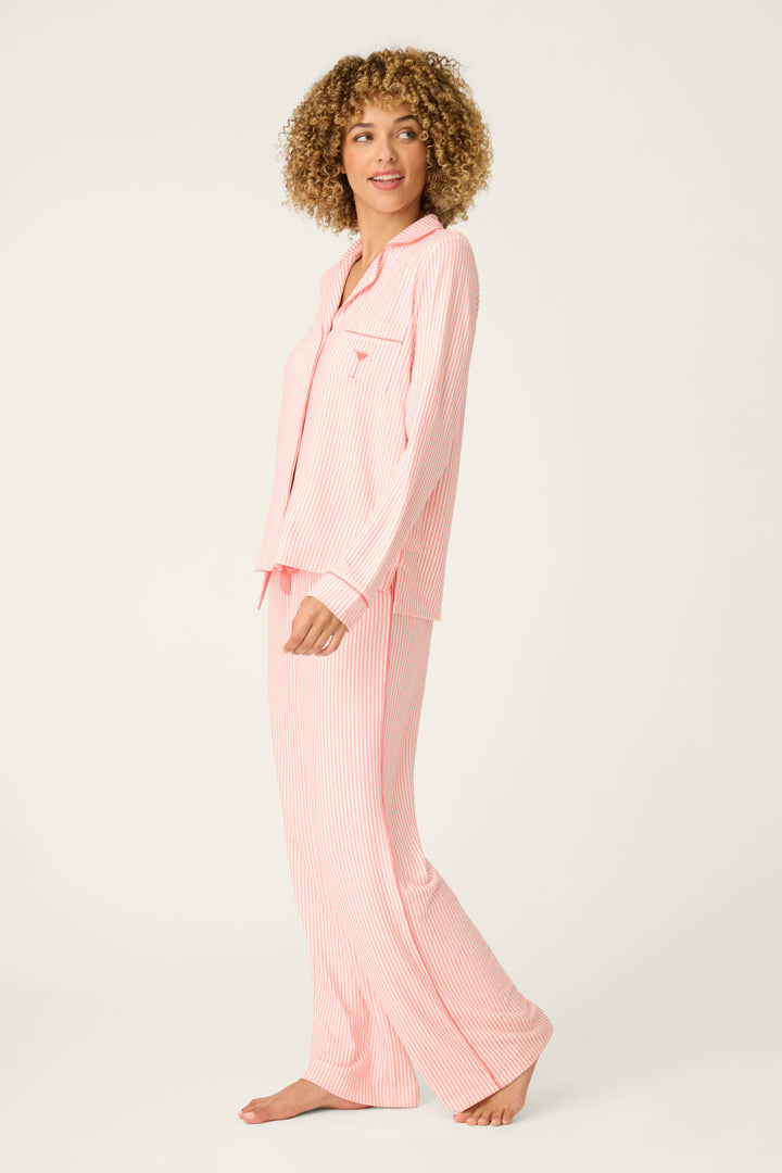 A person with curly hair stands barefoot against a plain light background, wearing the Paloma Mama Sweet Stripes Long 3-Piece PJ Set by P.J. Salvage, featuring grapefruit pink stripes and long sleeves with full-length pants.