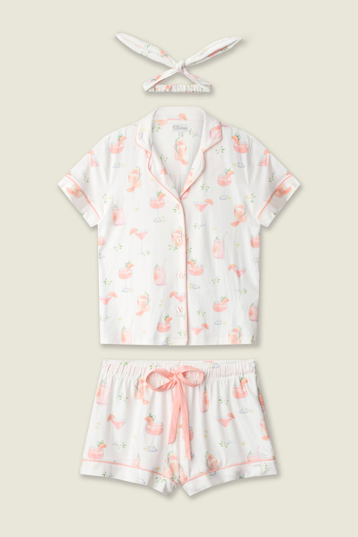 The P.J. Salvage Paloma Mama Sweet Sips 3-Piece PJ Set in butter jersey is laid flat, showcasing a short-sleeve button-up shirt, drawstring shorts, and headband with a playful pink flamingo and green leaf print.