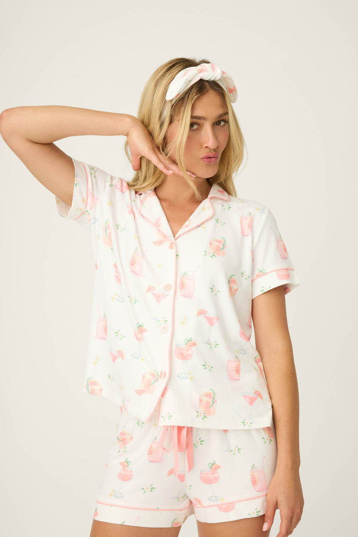 A woman with long blonde hair models the P.J. Salvage Paloma Mama Sweet Sips 3-Piece PJ Set, featuring a playful paloma cocktail print, and a white headband. She poses with one hand on her cheek against a plain light background.