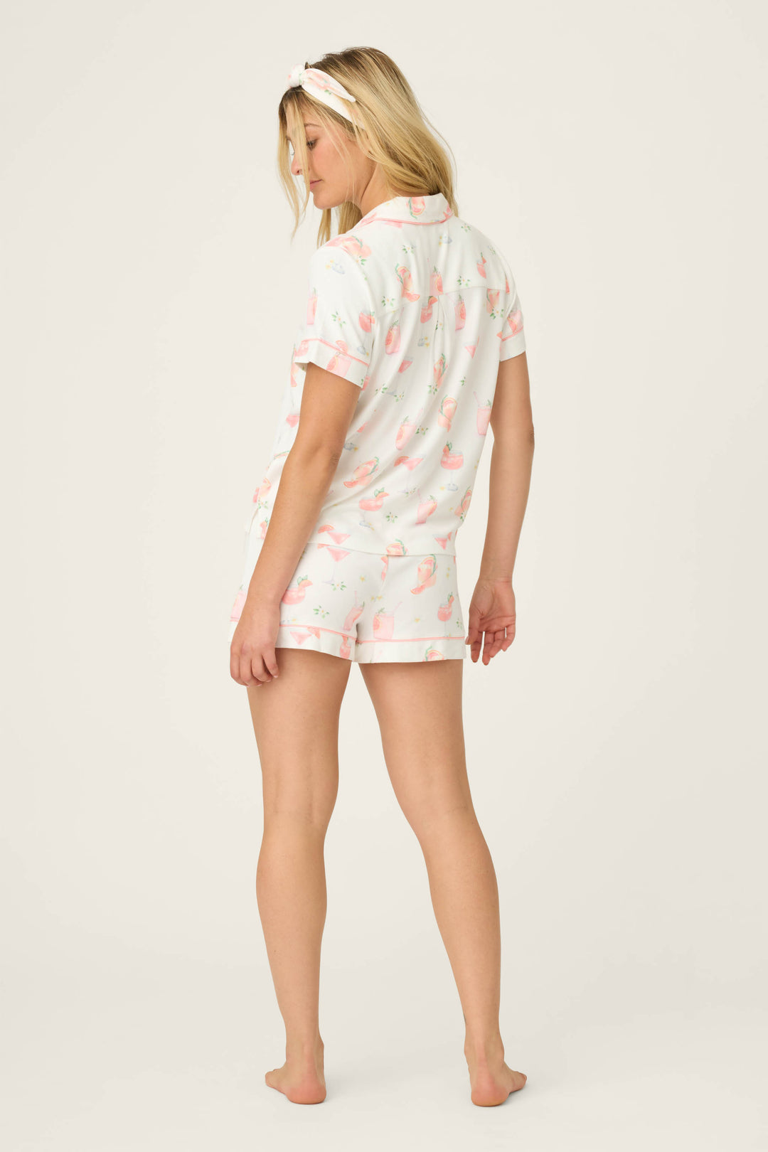 A woman stands barefoot on a plain background, facing away. She wears P.J. Salvage's Paloma Mama Sweet Sips 3-Piece PJ Set—matching butter jersey short-sleeve top, flamingo print shorts, and a coordinating headband.
