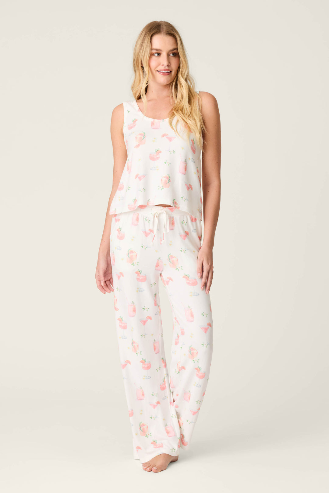 A woman with long blonde hair and a neutral expression stands barefoot in P.J. Salvage's Paloma Mama Sweet Sips Open Leg Pajama Pants, part of a sleeveless white butter jersey set with a playful Paloma cocktail print and matching ivory lounge pants.
