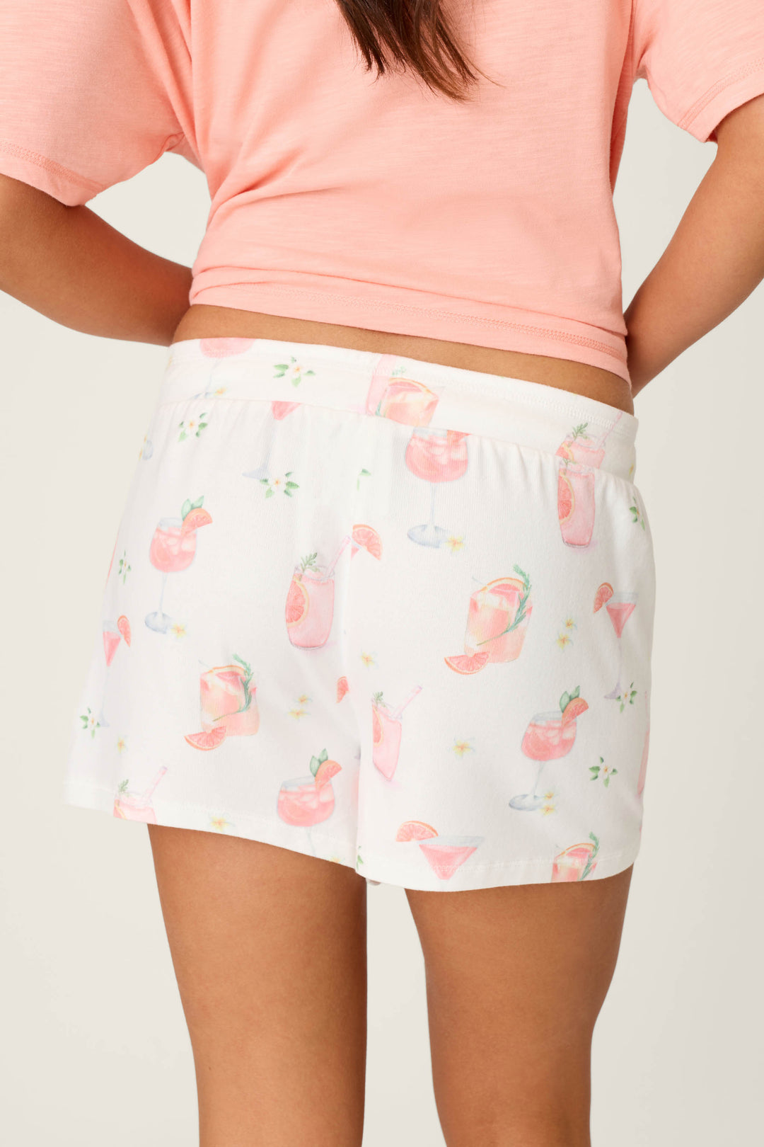 The image shows a person from the back, wearing P.J. Salvage Paloma Mama Sweet Sips Shorts—a relaxed fit lounge short—paired with a light pink shirt featuring a colorful cocktail drink print. Only the waist to upper thigh is visible.