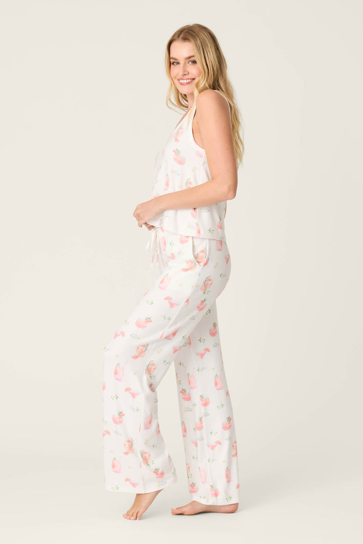 A woman with long blonde hair stands barefoot, wearing the Paloma Mama Sweet Sips Long PJ Set by P.J. Salvage, which features a pink and green floral pattern. She faces sideways, smiling slightly against a plain light background.