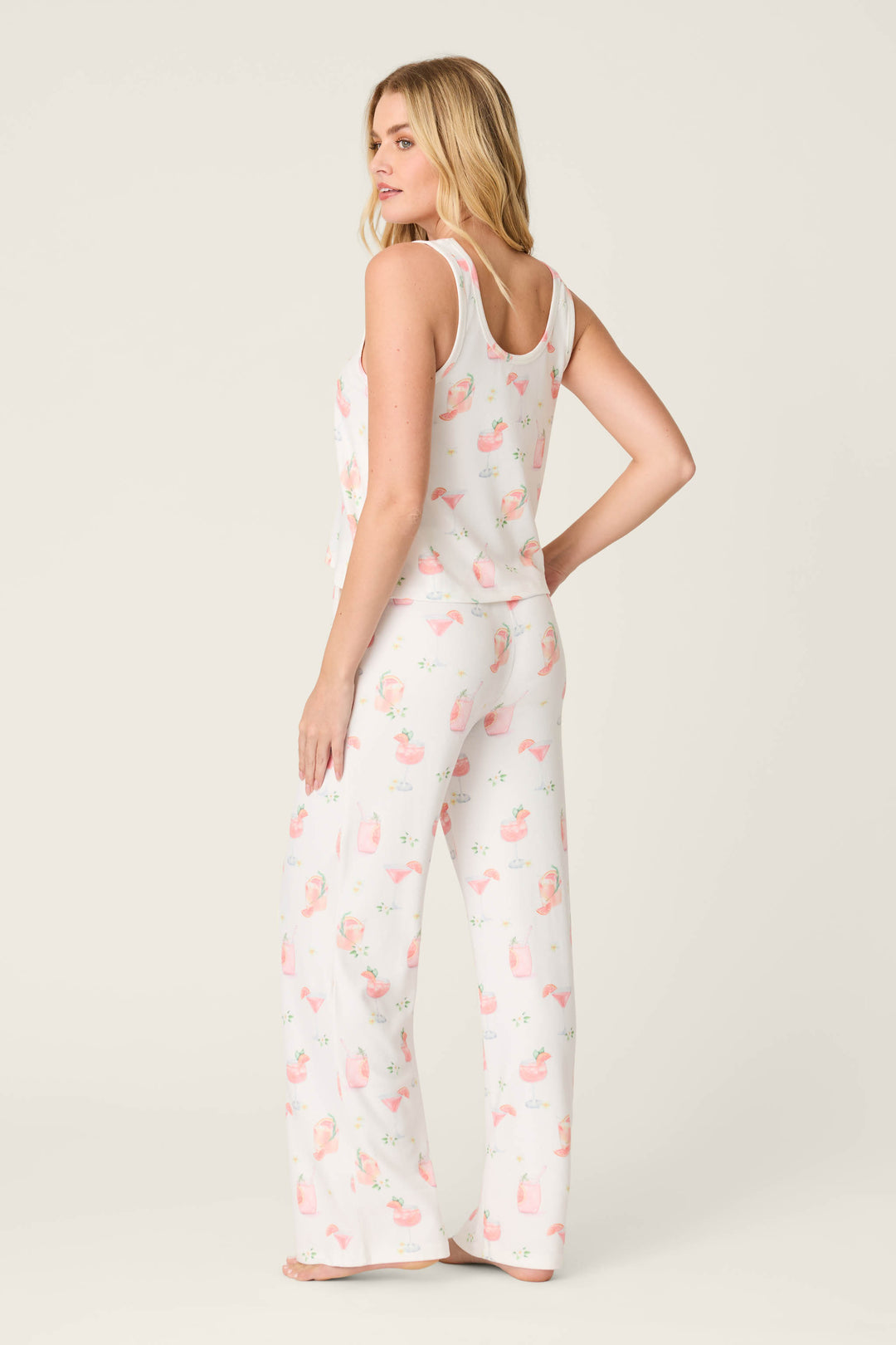 A woman stands barefoot on a plain background, modeling the P.J. Salvage Paloma Mama Sweet Sips Long PJ Set in ivory with a light pink and orange print. She faces away from the camera, turning her head to the side.