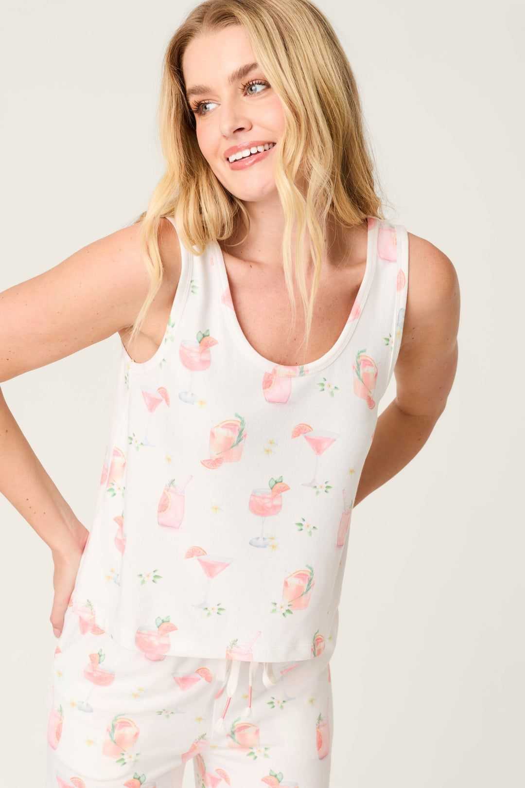 A woman with long blonde hair wears the P.J. Salvage Paloma Mama Sweet Sips Tank Top in breathable butter jersey, printed with pink flamingos and tropical drinks, smiling and looking slightly to the side against a light background.