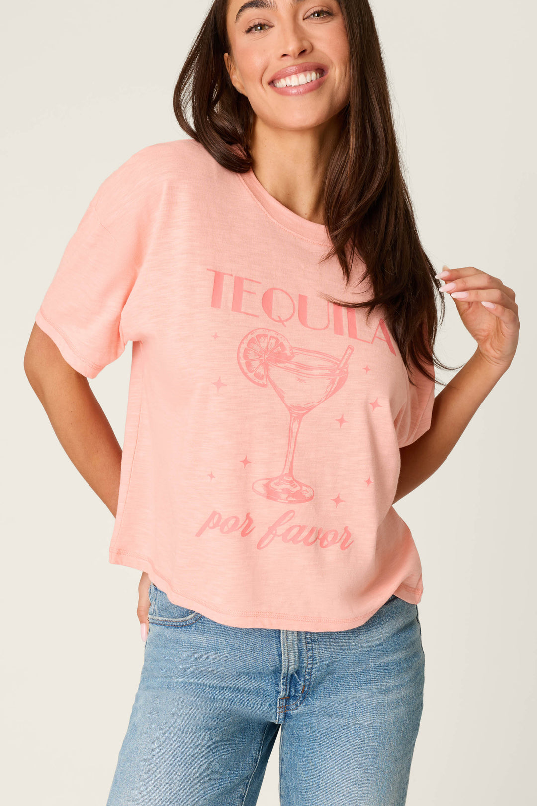 A woman in the P.J. Salvage Paloma Mama Tequila Por Favor Graphic T-Shirt, a grapefruit pink crew tee in soft cotton-modal slub jersey, smiles at the camera. She pairs it with light blue jeans for a comfortable look.