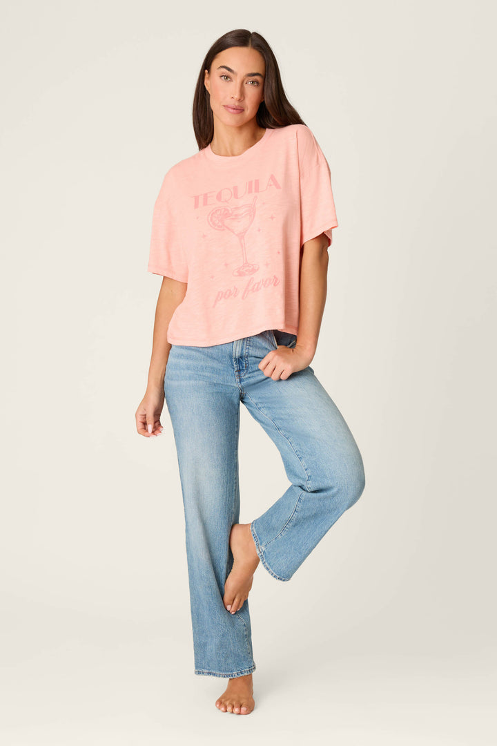 A woman stands barefoot on one leg, wearing light blue jeans and the P.J. Salvage Paloma Mama Tequila Por Favor Graphic T-Shirt in grapefruit pink cotton-modal slub jersey, featuring a playful margarita design, against a plain light background.