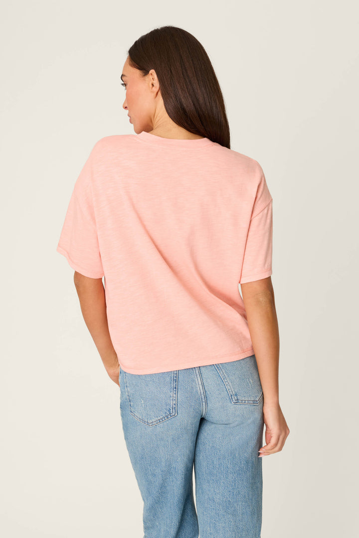 A person stands facing away from the camera, wearing the P.J. Salvage Paloma Mama Tequila Por Favor Graphic T-Shirt and light blue jeans against a plain, light background.