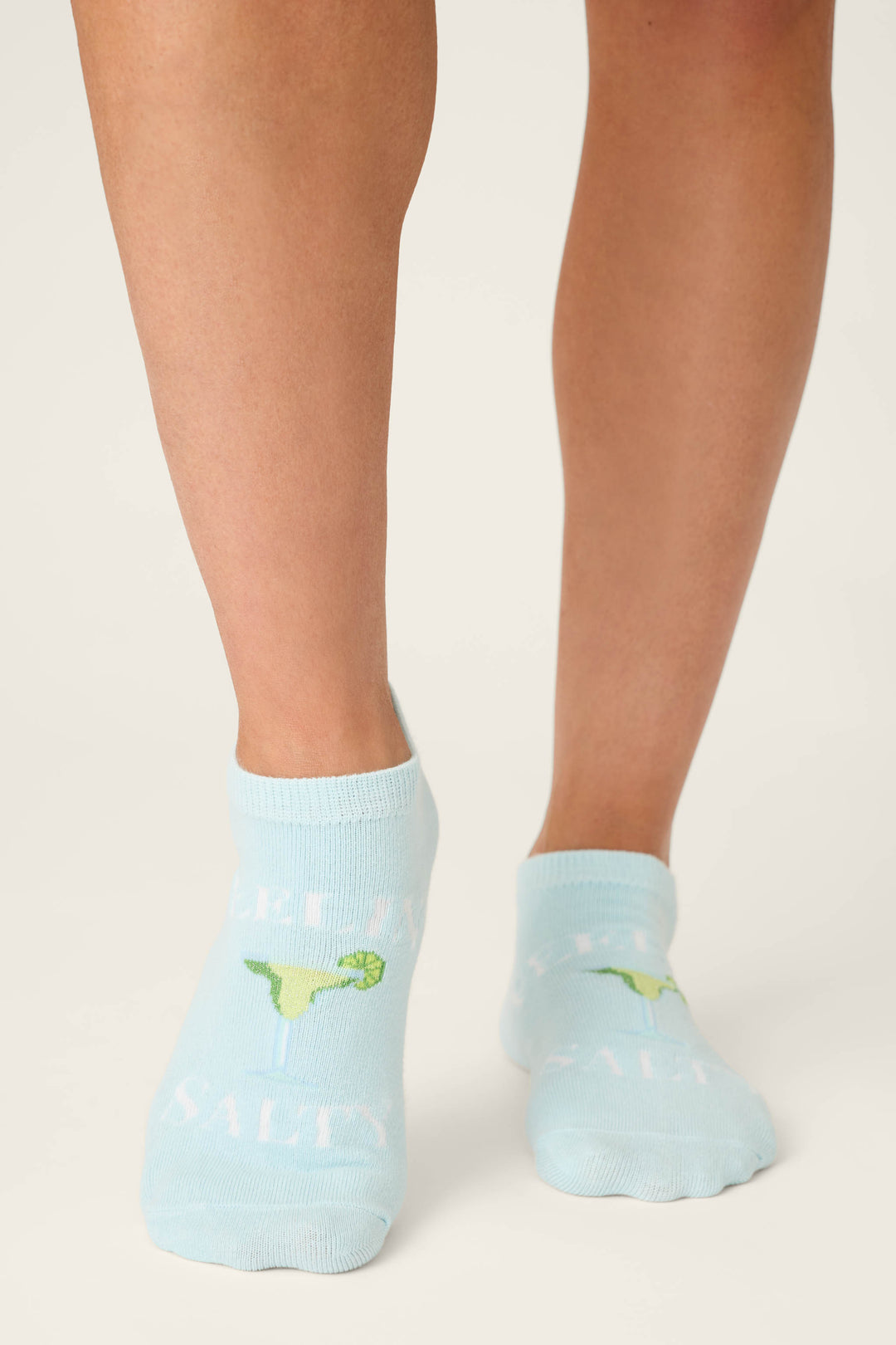 A person wears P.J. Salvage Playful Prints Feeling Salty 3-Pack No Show Socks in light blue with grip soles and a green dinosaur graphic, visible from mid-calf down against a neutral background.