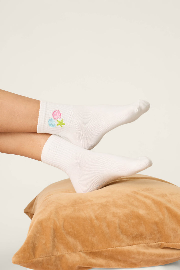 A person wears P.J. Salvage Playful Prints She Sells Seashells Quarter Crew Socks, featuring small pastel seashell and starfish designs, while resting their feet on a tan velvet pillow against a plain background.