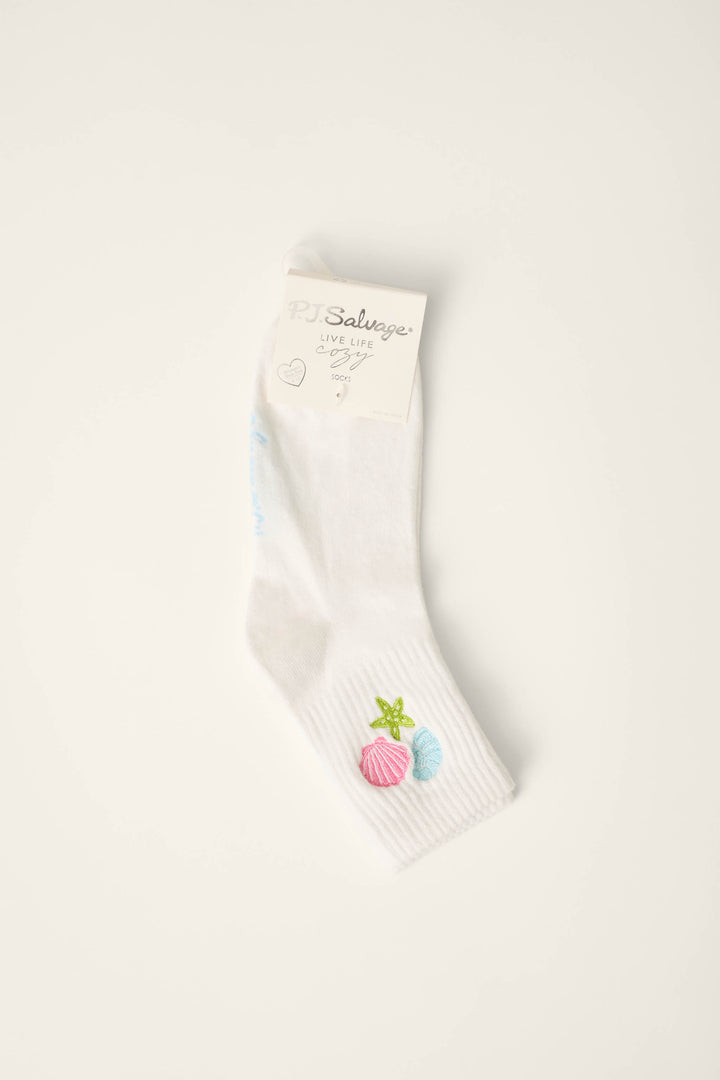 The Playful Prints She Sells Seashells Quarter Crew Socks by P.J. Salvage are white with a “P.J. Salvage” label and feature embroidered pink shells, a green starfish, and a blue shell near the ankle.