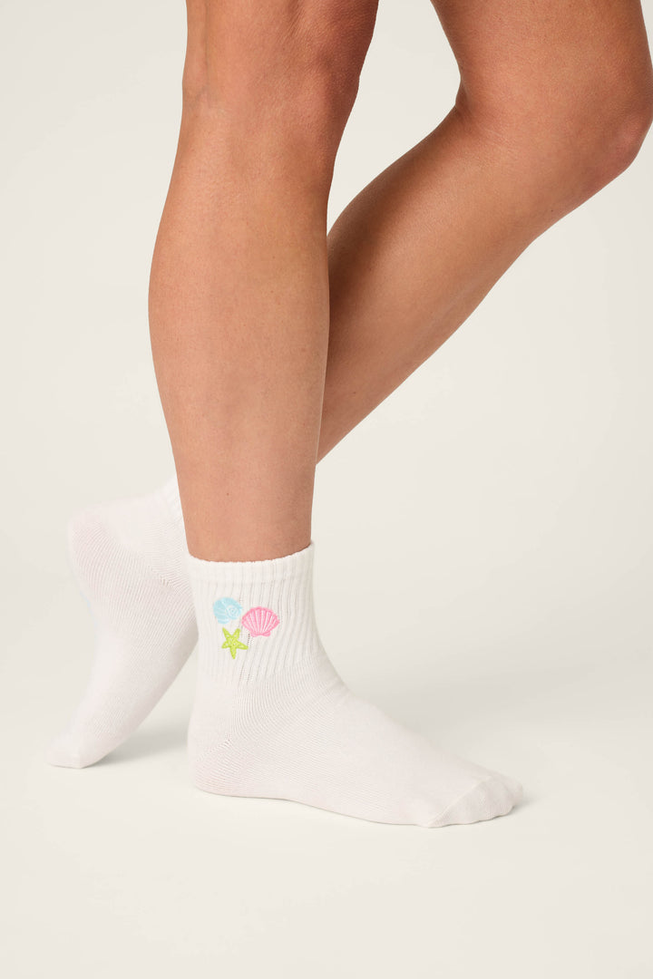 A person models P.J. Salvage's Playful Prints She Sells Seashells Quarter Crew Socks, featuring small pastel shell and starfish designs, standing with one foot slightly behind the other against a plain backdrop.