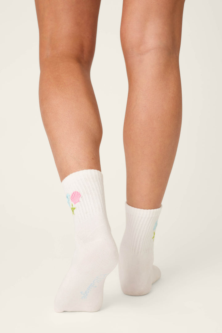 A person is seen from behind, showing their lower legs and feet in P.J. Salvage Playful Prints She Sells Seashells Quarter Crew Socks with small colorful designs like a pink shell and green star, standing on a light background.