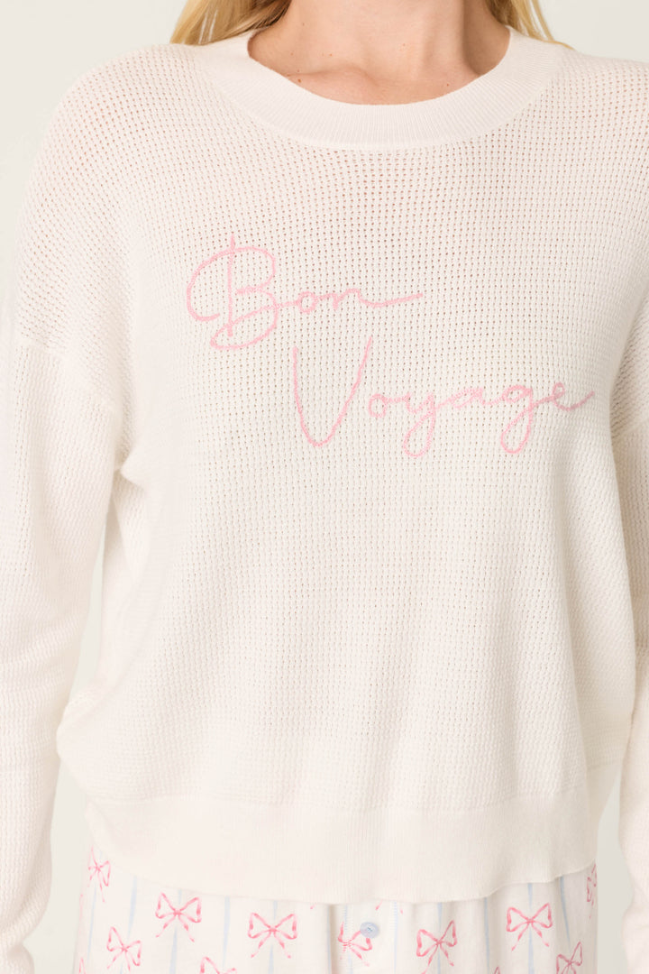 A person wears the P.J. Salvage Playful Prints Bon Voyage Pullover Sweater in ivory with pink script, along with pajama pants featuring pink bows.