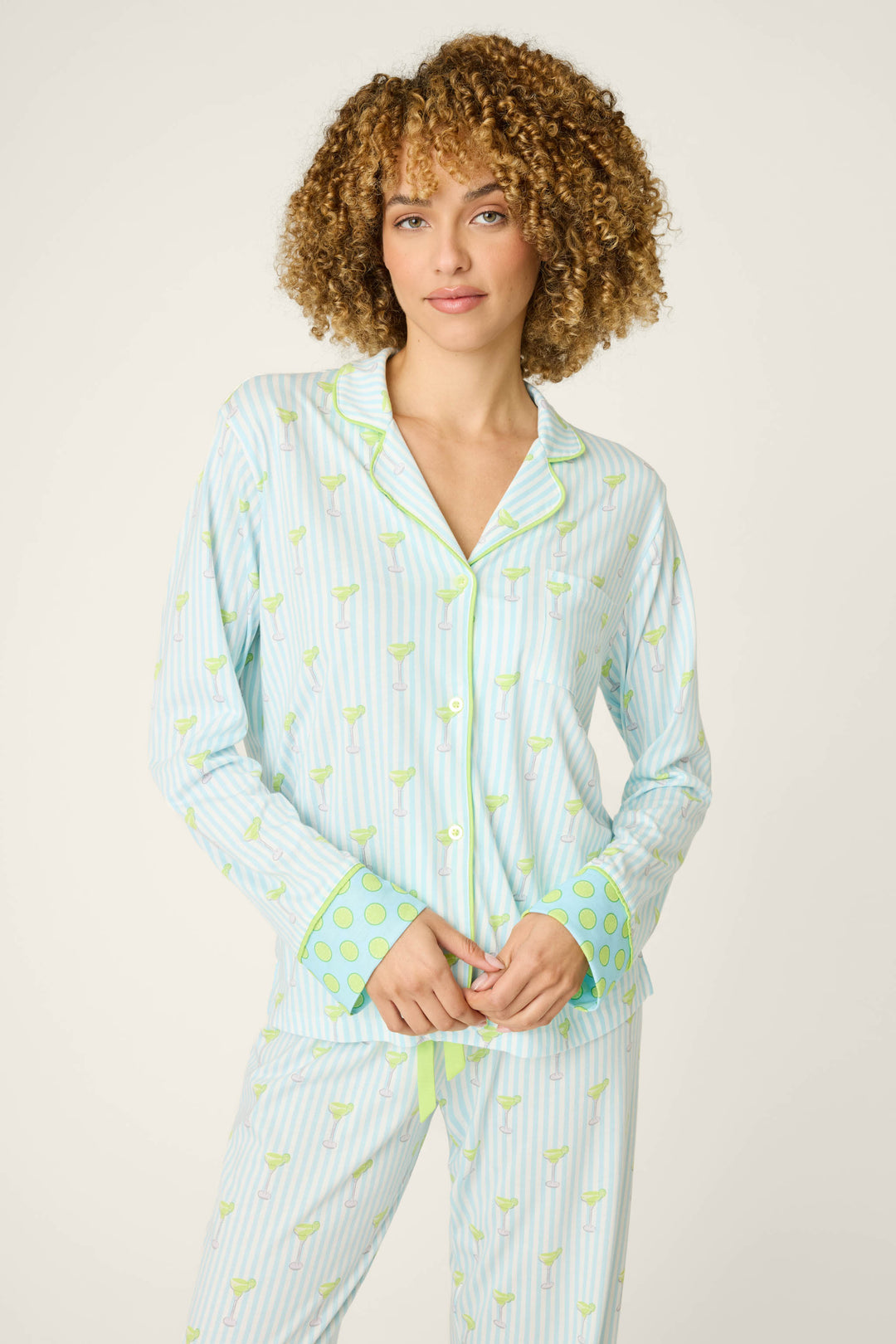 A person with curly hair wears the P.J. Salvage Playful Prints Salty Cadillac Long Sleeve Top, a soft cotton-modal jersey pajama shirt with light blue and white stripes, green patterns, and polka-dot cuffs, against an off-white background.