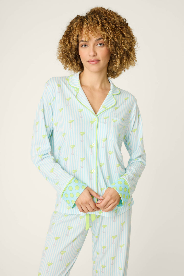 A person with curly hair wears the P.J. Salvage Playful Prints Salty Cadillac Long Sleeve Top, a soft cotton-modal jersey pajama shirt with light blue and white stripes, green patterns, and polka-dot cuffs, against an off-white background.