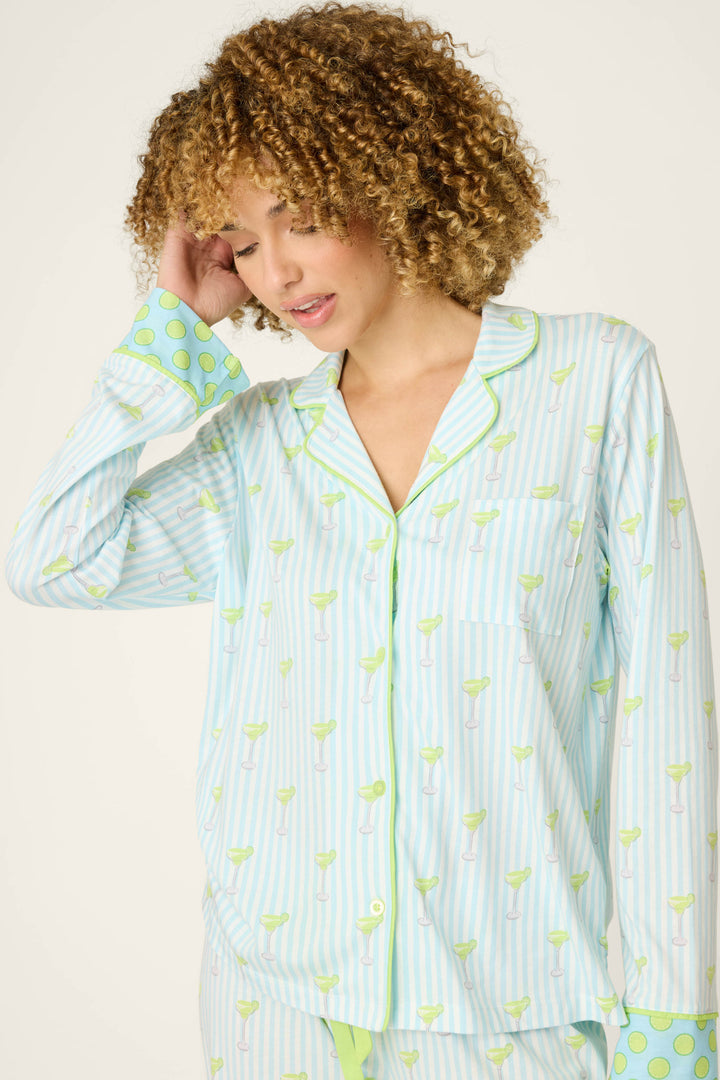 A woman with curly hair wears the P.J. Salvage Playful Prints Salty Cadillac Long Sleeve Top—light blue and white striped sleepwear with green margarita motifs and polka dot cuffs—looking down as she gently touches her hair.