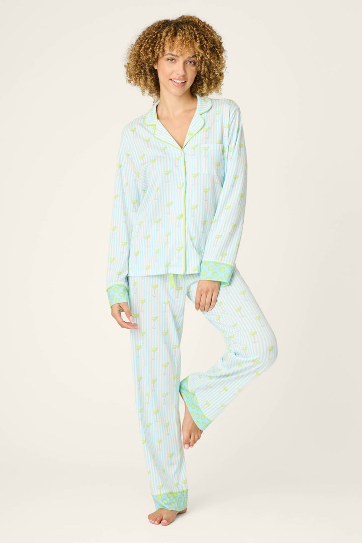 A woman with curly hair wears the P.J. Salvage Playful Prints Salty Cadillac Long Sleeve Top, a striped light blue and white cotton-modal jersey with green accents. She stands barefoot, smiling at the camera against a plain background.