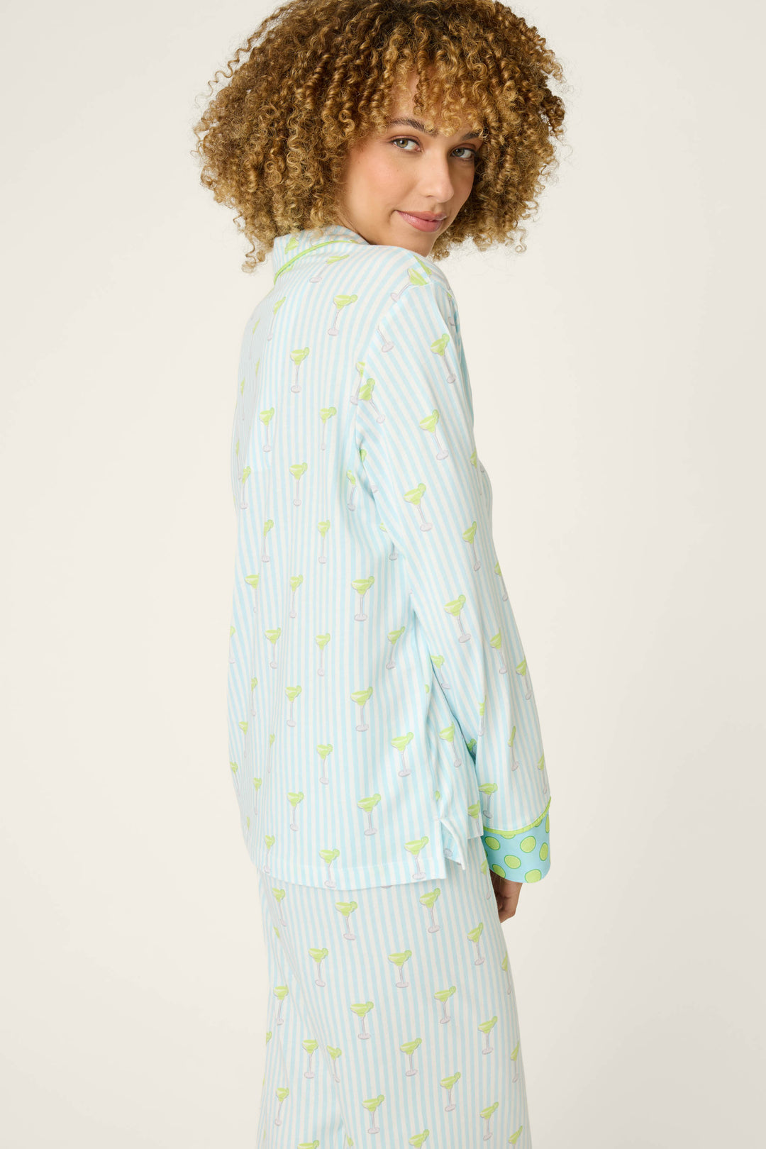 A person with curly hair wears the P.J. Salvage Playful Prints Salty Cadillac Long Sleeve Top, a light blue and white striped pajama shirt with a green pattern, standing sideways and looking over their shoulder at the camera against a plain, light background.