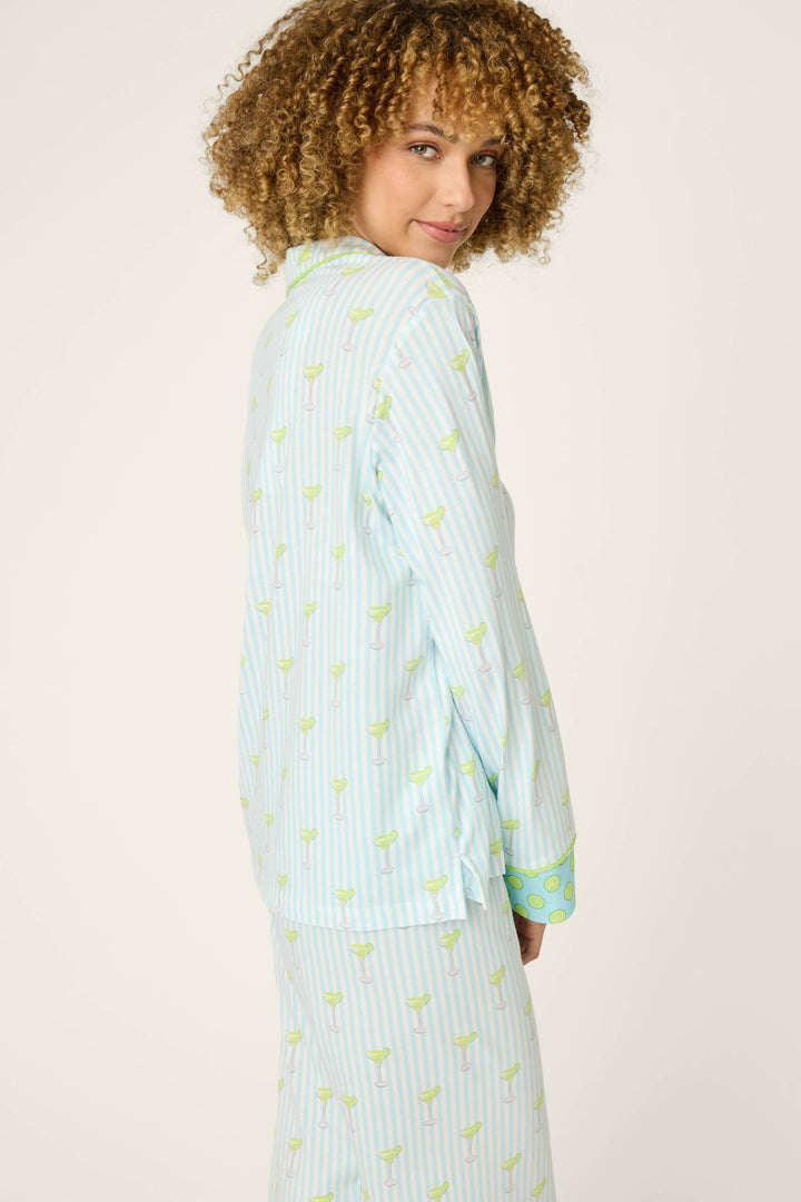 A person with curly hair wears the P.J. Salvage Playful Prints Salty Cadillac Long Sleeve Top, a light blue and white striped pajama shirt with a green pattern, standing sideways and looking over their shoulder at the camera against a plain, light background.