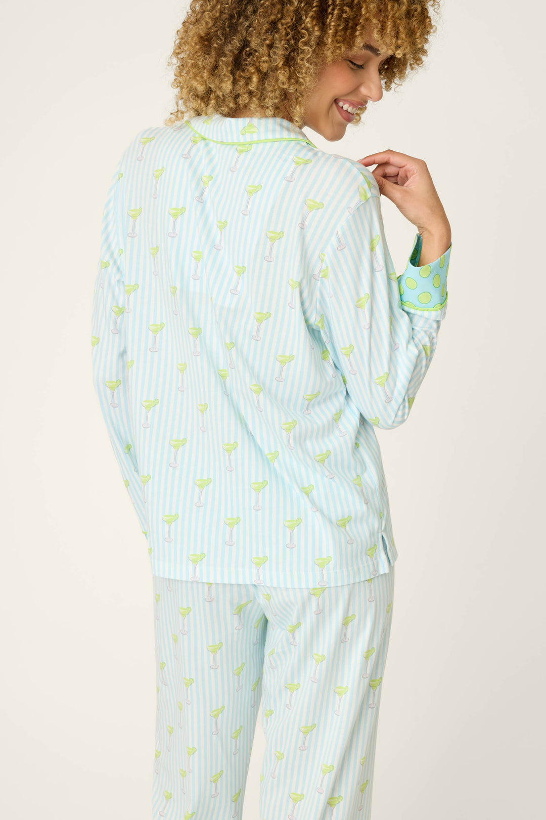 A person with curly hair, shown from the back and smiling, wears the P.J. Salvage Playful Prints Salty Cadillac Long Sleeve Top—light blue and white striped sleepwear with green margarita motifs and green polka dot cuffs.