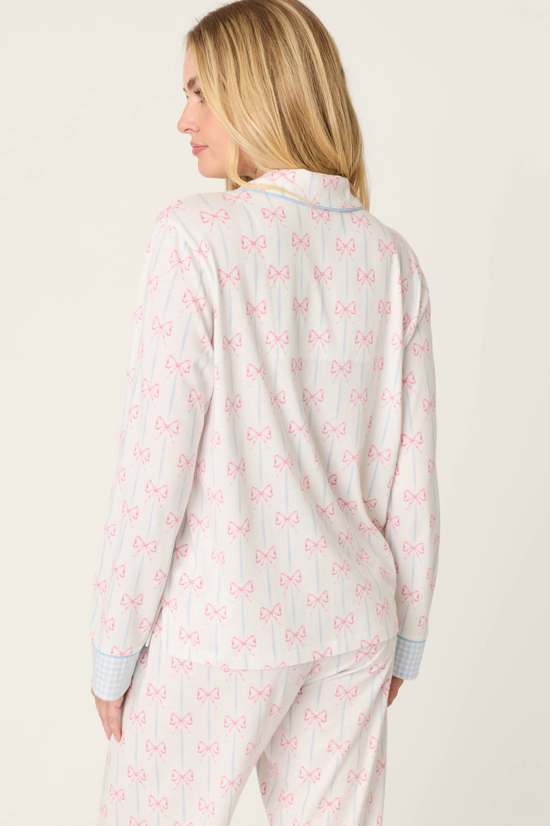 A woman with long blonde hair stands against a light background wearing the P.J. Salvage Playful Prints All Wrapped Up Long Sleeve Top—white cotton-modal jersey pajamas with pink bows and blue gingham cuffs.