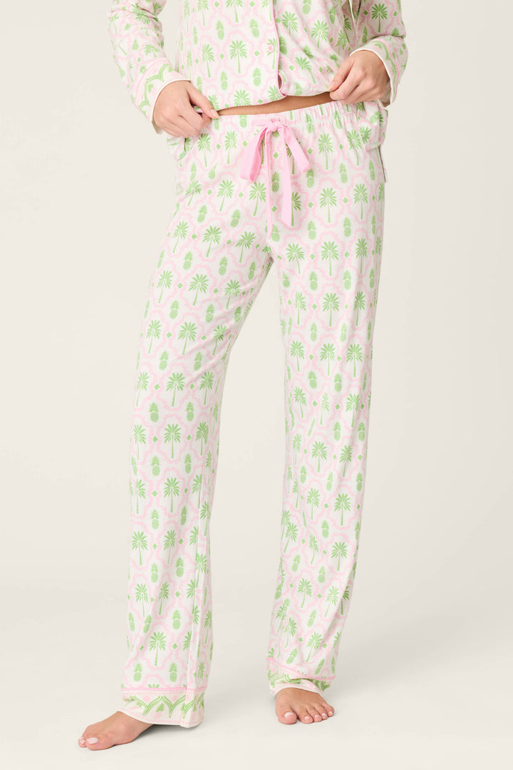 A person models the Playful Prints Island Time Open Leg PJ Pants by P.J. Salvage—light pink with green palm trees and a pink drawstring, in soft cotton-modal jersey—standing barefoot on a white background with the matching pajama top partly visible.
