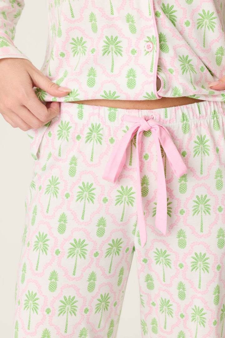 A person wears P.J. Salvage’s Playful Prints Island Time Open Leg PJ Pants—light pink and white with a green palm tree and pinecone print—paired with a partially tucked top and tied at the waist with a pink drawstring.