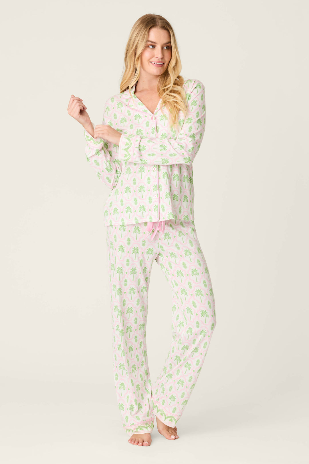 A person stands barefoot in P.J. Salvage Playful Prints Island Time Open Leg PJ Pants, featuring a green and pink palm tree pattern, looking to the side and touching one sleeve against a plain light background.