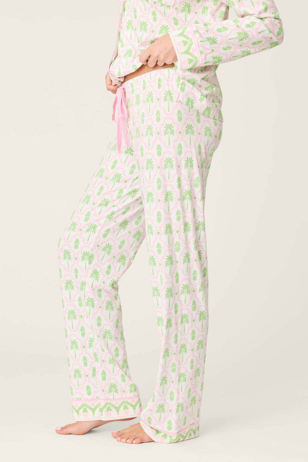 A person stands barefoot, wearing P.J. Salvage Playful Prints Island Time Open Leg PJ Pants in light pink and white with a green palm tree pattern and pink drawstring. The matching pajama top is partially visible.