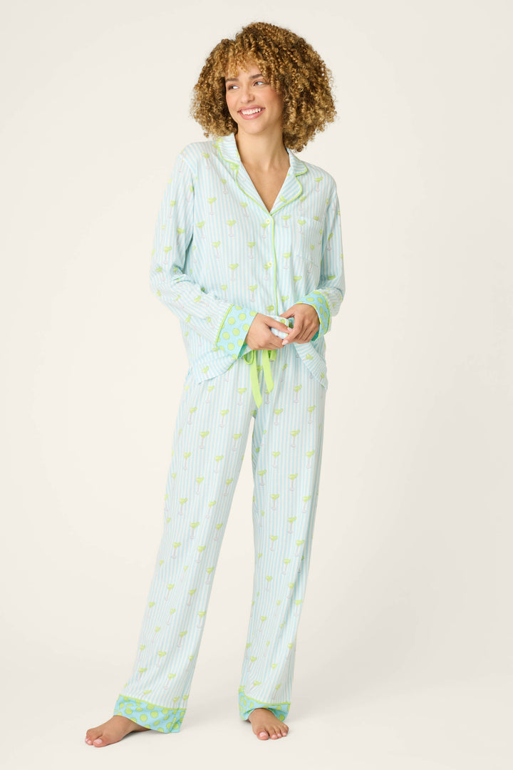 A person with curly hair stands barefoot against a plain background in the P.J. Salvage Playful Prints Island Time Short PJ Set, featuring light blue and white stripes, small yellow prints, green trim at the cuffs and hems, and a green drawstring waist.