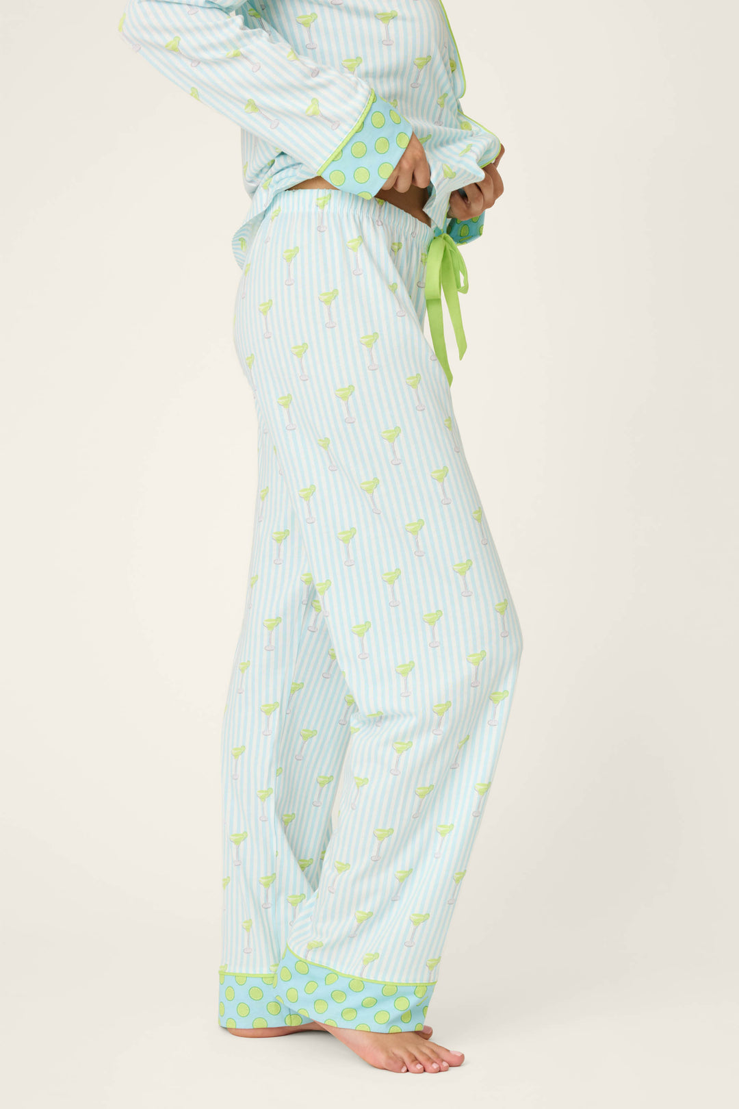 A person is wearing P.J. Salvage’s Playful Prints Island Time Short PJ Set pajama pants in light blue and white stripes, with a green drawstring, green polka dot cuffs, and small green objects. They’re barefoot, upper body partially out of frame.