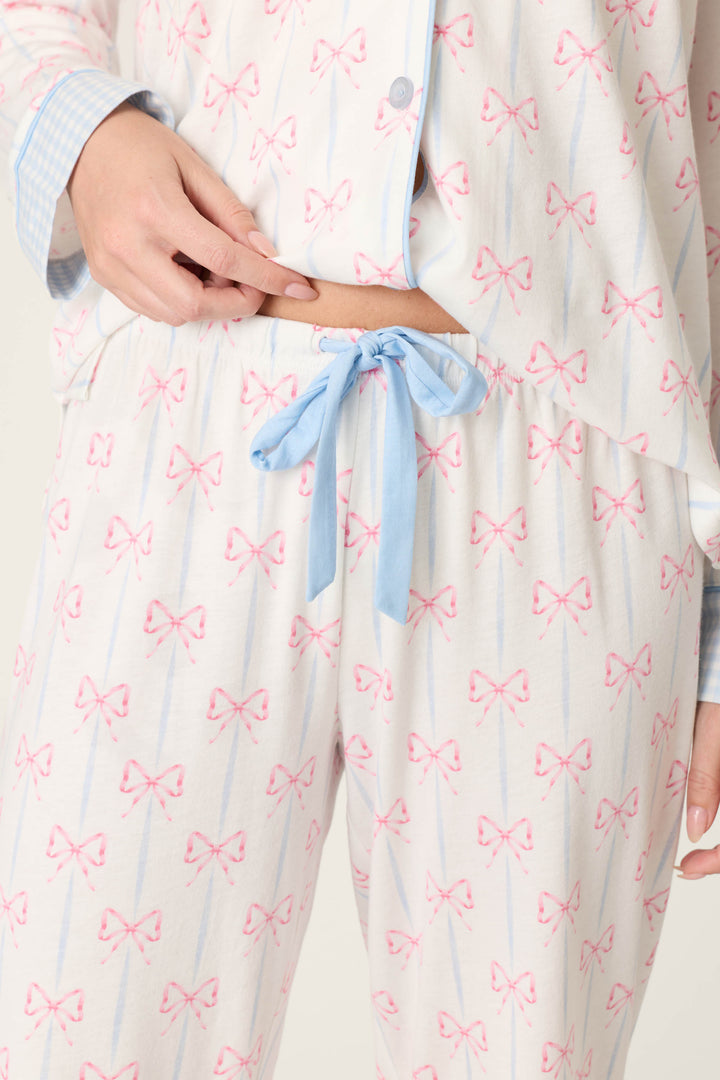 A person wears P.J. Salvage's Playful Prints All Wrapped Up Open Leg PJ Pants, featuring a white bow print and blue drawstring waist, holding the waistband while the relaxed open-leg silhouette is visible beneath a partially unbuttoned pajama top.