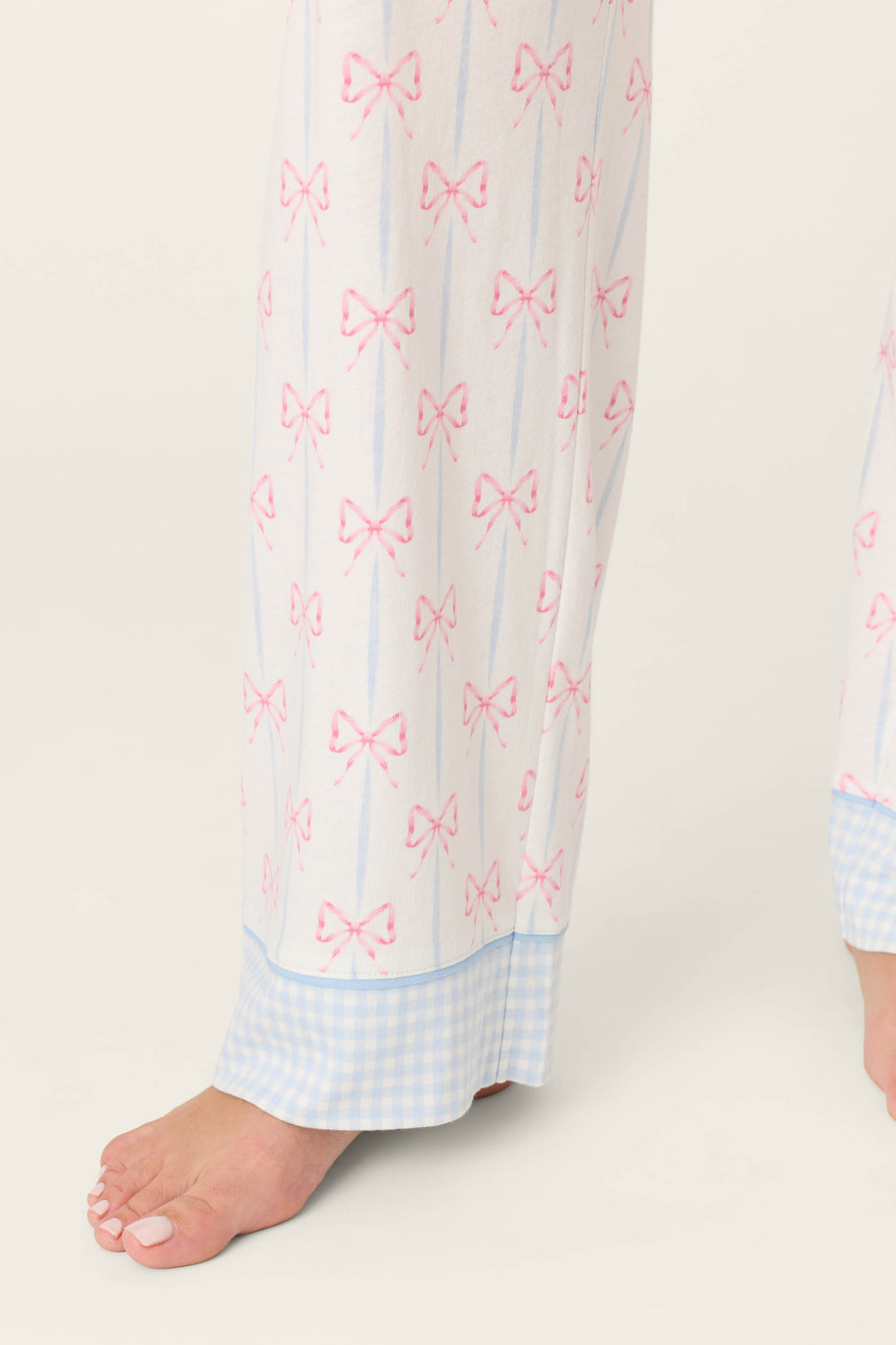 A person models the P.J. Salvage Playful Prints All Wrapped Up Open Leg PJ Pants, featuring bow prints and blue gingham cuffs, standing barefoot on a light background with only their lower legs and feet visible.