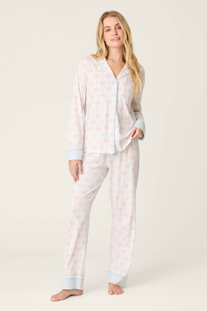 A woman stands barefoot against a plain background, wearing P.J. Salvage's Playful Prints All Wrapped Up Open Leg PJ Pants—bow-print pants with blue plaid cuff trim and a comfy drawstring waist—paired with a matching long-sleeve button-up top.