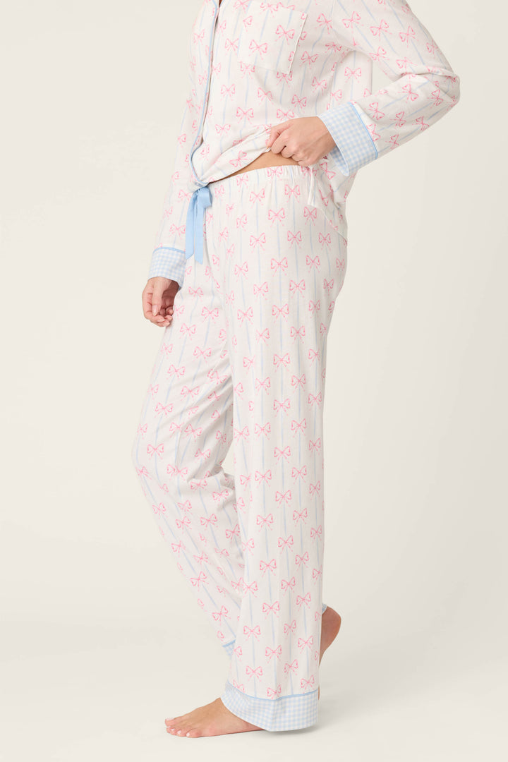 A person stands barefoot against a plain background, showing only the lower half in P.J. Salvage Playful Prints All Wrapped Up Open Leg PJ Pants featuring a bow print, blue checkered cuffs, and an open-leg silhouette.