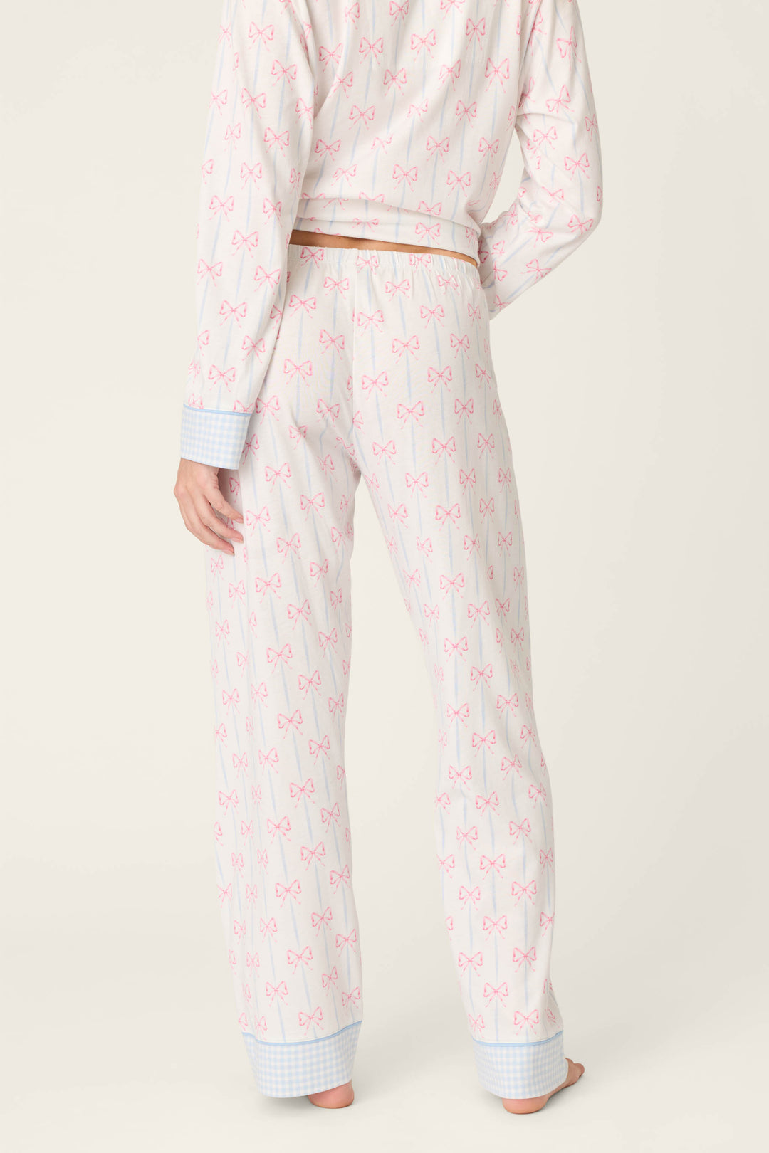 A person stands barefoot facing away, wearing P.J. Salvage’s Playful Prints All Wrapped Up Open Leg PJ Pants with a bow print, drawstring waist, blue checkered cuffs, and open-leg silhouette, set against a plain light beige background.