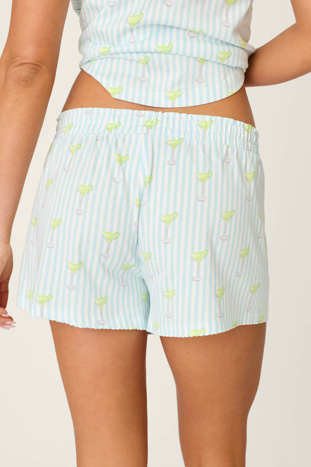 A person wears P.J. Salvage Playful Prints Salty Cadillac PJ Shorts, featuring a blue-and-white stripe and margarita glass print in soft cotton-modal jersey. The photo highlights the back of the shorts and the wearer's lower back.