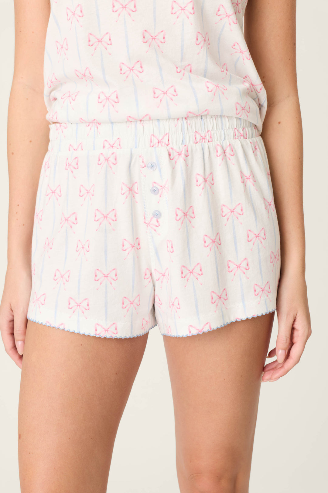 A person wears P.J. Salvage Playful Prints All Wrapped Up PJ Shorts, featuring bow prints, blue buttons, and scalloped picot hems, paired with a matching soft cotton-modal top. The photo is cropped from mid-torso to mid-thigh.