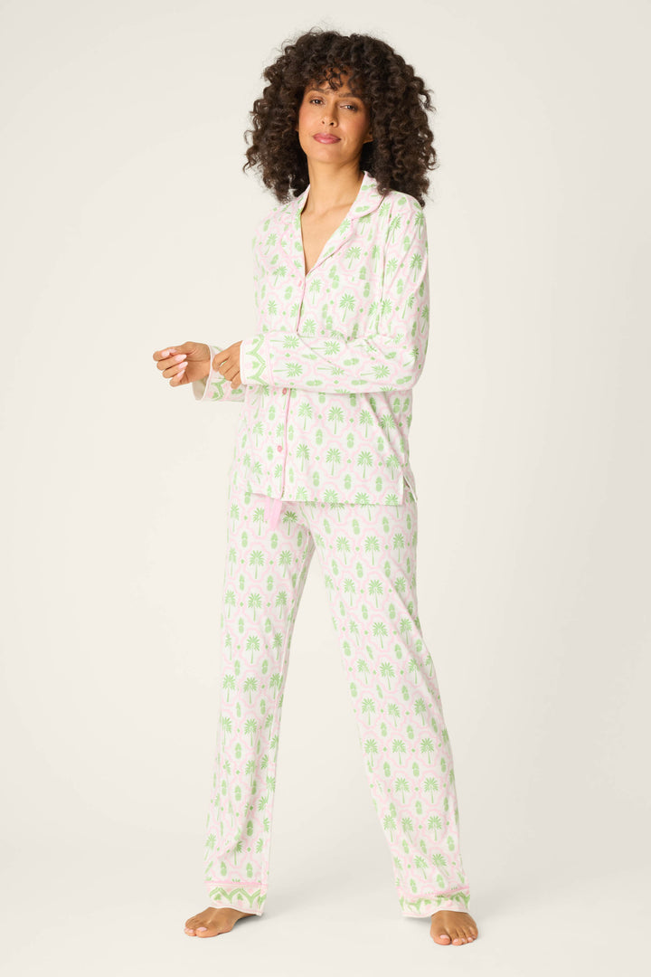 A person with curly hair stands barefoot, wearing the P.J. Salvage Playful Prints Island Time Long PJ Set in a light pink and green pattern on white, made from soft cotton-modal jersey, against an off-white background.