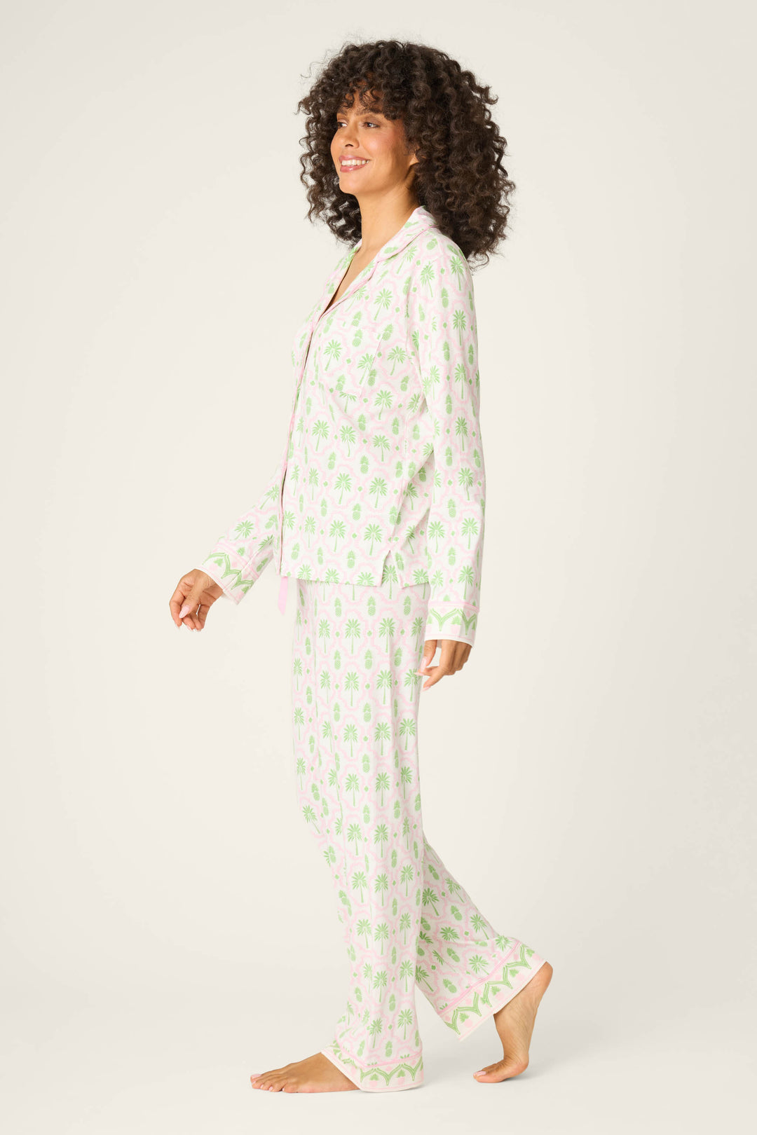 A person stands sideways, smiling and barefoot, in the P.J. Salvage Playful Prints Island Time Long PJ Set—an ivory cotton-modal jersey pajama set with a light green palm tree print, button-up top, and long pants against a white background.