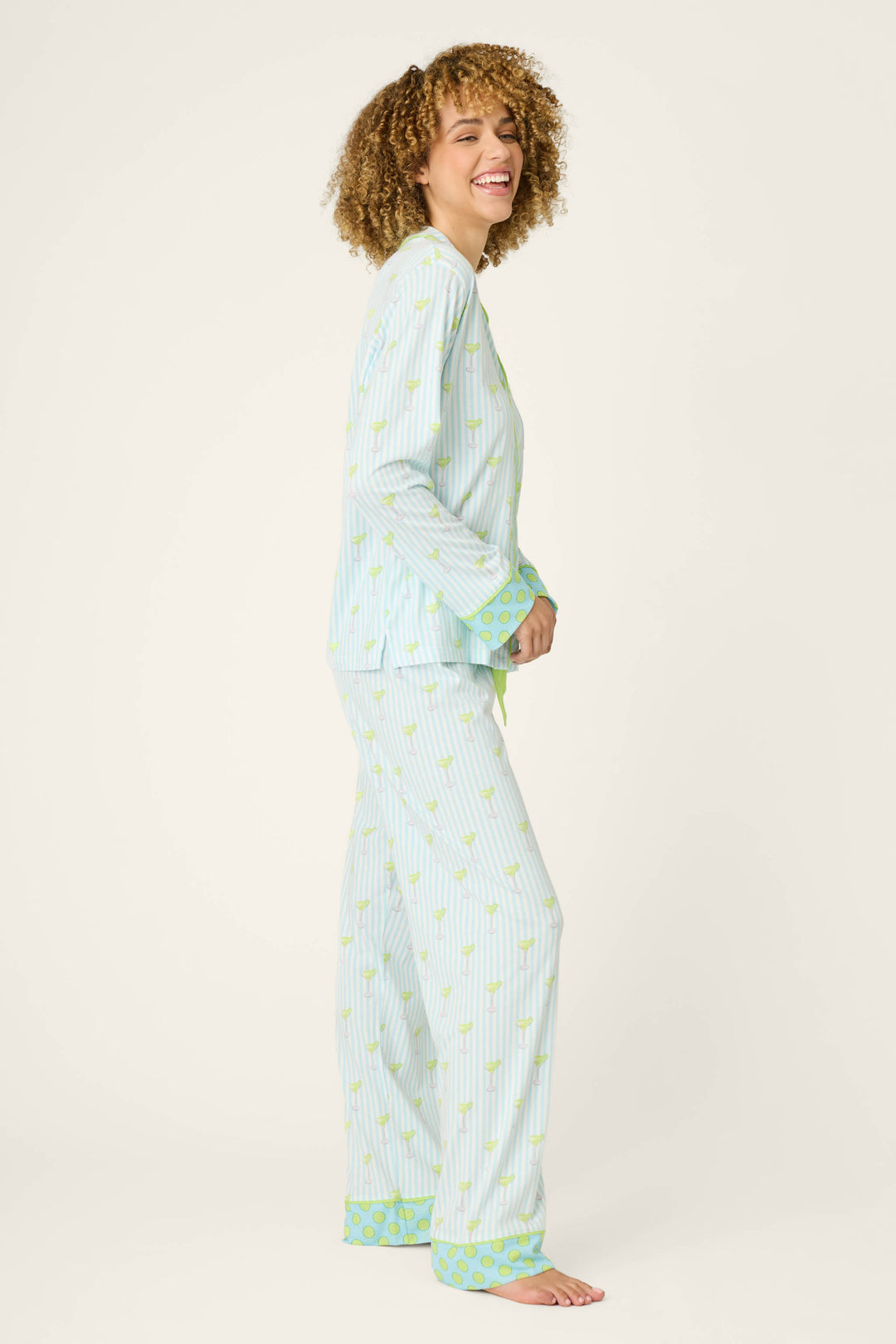 A person with curly hair stands sideways, smiling, wearing the P.J. Salvage Playful Prints Salty Cadillac Striped Long PJ Set—cotton-modal jersey with a small yellow print and green patterned cuffs—against a plain light background.