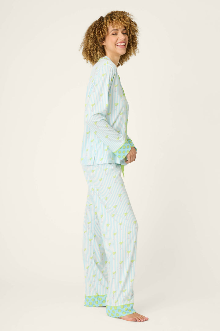 A person with curly hair stands sideways, smiling, wearing the P.J. Salvage Playful Prints Salty Cadillac Striped Long PJ Set—cotton-modal jersey with a small yellow print and green patterned cuffs—against a plain light background.