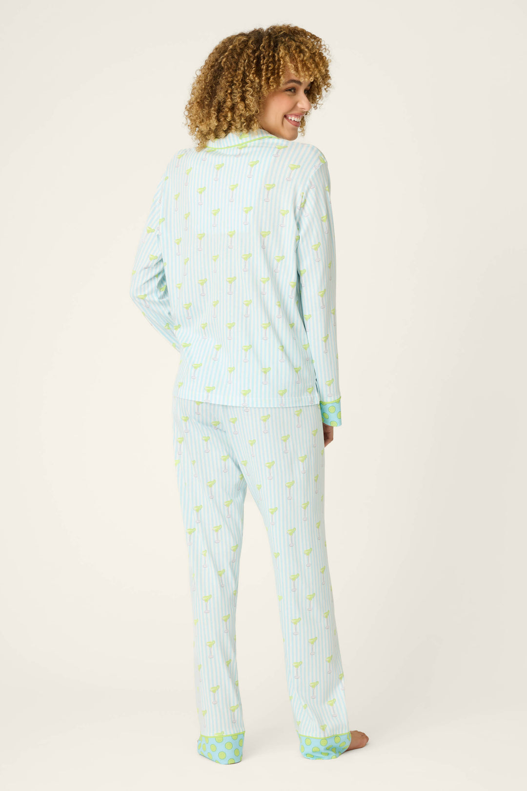 Model stands barefoot, smiling over their shoulder with curly hair, in the P.J. Salvage Playful Prints Salty Cadillac Striped Long PJ Set—featuring a fun pineapple print and green accents in soft cotton-modal jersey—against a plain background.