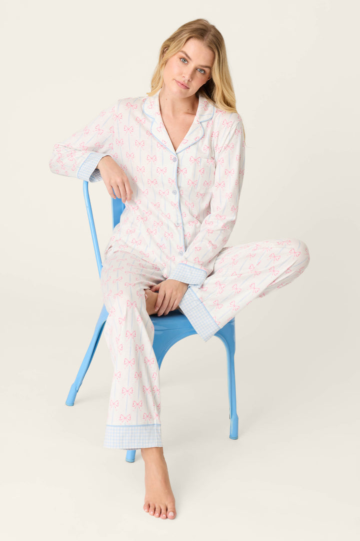 A woman with long blonde hair sits barefoot on a blue chair, wearing the Playful Prints All Wrapped Up Long PJ Set by P.J. Salvage, featuring a pink bow-print pattern and light blue checkered accents, against a plain light background.