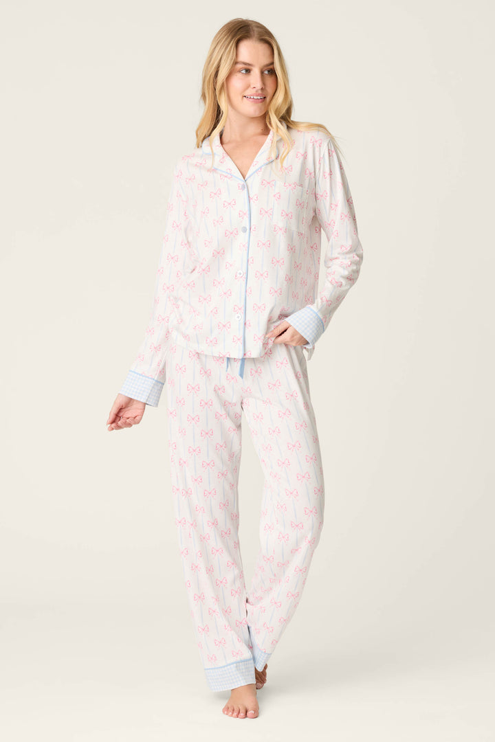 A woman with long blonde hair stands barefoot against a plain background, wearing the P.J. Salvage Playful Prints All Wrapped Up Long PJ Set—white with pink bows, and a button-front shirt featuring blue cuffs and buttons.